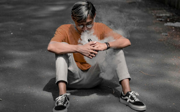 a man with glasses, orange t-shirt and khaki pants sitting on concrete pavement while vaping