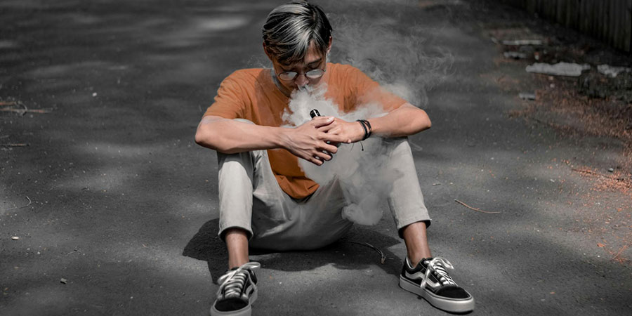 a man with glasses, orange t-shirt and khaki pants sitting on concrete pavement while vaping