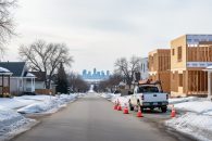 Late-winter Saskatoon residential street with older bungalows, a new infill home under construction, orange roadwork cones, and an unbranded utility truck, with bare trees, snowbanks, and a faint city skyline in the distance.