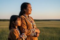 Indigenous mother and child standing on the Saskatchewan prairie during golden hour with a soft-focus treeline in the background.