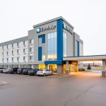 Exterior view of the Saskatoon Travelodge on Circle Drive West with driveway and parking area under a bright overcast sky.